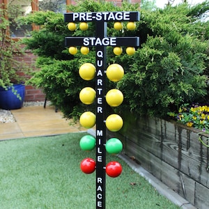 Op de afbeelding: Een zwarte metalen kerstboomstandaard met een zwarte basis en een verticale paal. De paal heeft een reeks gele, groene en rode ballen eraan bevestigd. De ballen zijn gelabeld met "Pre-Stage", "Stage", "Quarter Mile Raceway".