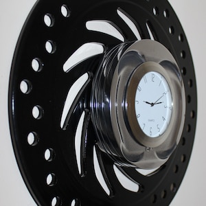 Op de afbeelding: Een zwarte metalen klok met een cirkelvormig ontwerp en een zilveren wijzerplaat. De klok is aan de muur gemonteerd en heeft een quartz uurwerk.