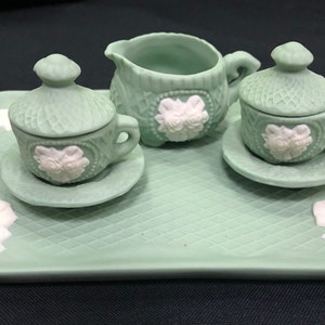 May include: A green ceramic tea set with a teapot, creamer, sugar bowl, and two teacups and saucers. The set is decorated with white roses and has a woven pattern.