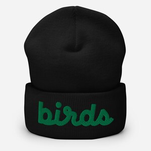 May include: Black beanie hat with the word "birds" embroidered in green. The hat has a folded cuff and a soft, knitted texture. The green embroidery provides a striking contrast against the black fabric. A stylish winter accessory.