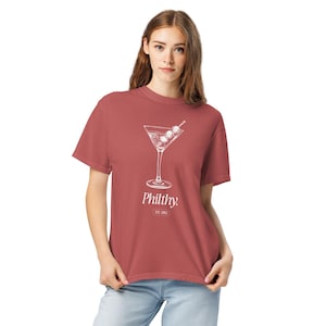 May include: A dusty rose t-shirt featuring a white graphic of a martini glass with olives. Below the graphic is the word "Philthy" and the text "EST. 1983". The shirt is a casual style.