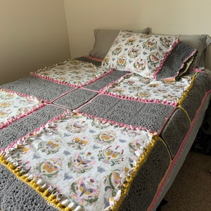 May include: A gray and white crocheted blanket with a butterfly pattern. The blanket is trimmed with pink and yellow crocheted edging.
