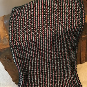 Op de afbeelding: Een gestreept gebreid deken in tinten donkerblauw, roze en grijs. Het deken heeft een textuur, een hobbelige oppervlak.