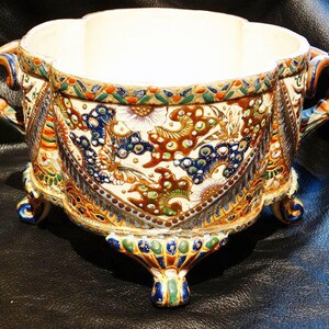 K&ouml;nnte beinhalten: Ein wei&szlig;er Keramiktopf mit einem gewellten Rand und drei Beinen. Der Topf ist mit einem bunten Blumenmuster in Blaut&ouml;nen, Gr&uuml;n, Gold und Braun verziert.