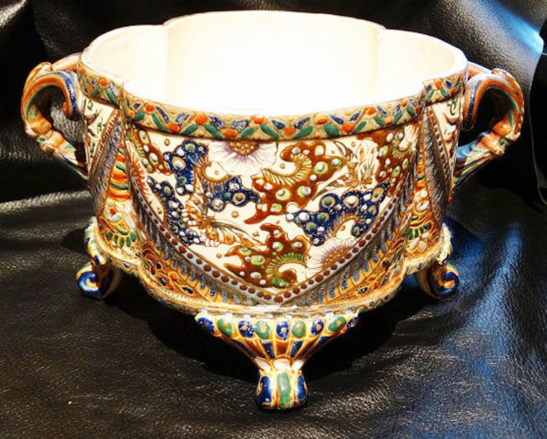 K&ouml;nnte beinhalten: Ein wei&szlig;er Keramiktopf mit einem gewellten Rand und drei Beinen. Der Topf ist mit einem bunten Blumenmuster in Blaut&ouml;nen, Gr&uuml;n, Gold und Braun verziert.
