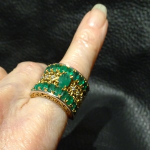 May include: A wide gold band ring with a row of emerald green gemstones set in a decorative filigree design.