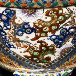 K&ouml;nnte beinhalten: Ein wei&szlig;er Keramiktopf mit einem detaillierten Blumen- und Schmetterlingsdesign. Der Topf ist mit blauen, gr&uuml;nen, goldenen und braunen Akzenten verziert.