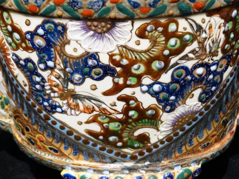 K&ouml;nnte beinhalten: Ein wei&szlig;er Keramiktopf mit einem detaillierten Blumen- und Schmetterlingsdesign. Der Topf ist mit blauen, gr&uuml;nen, goldenen und braunen Akzenten verziert.