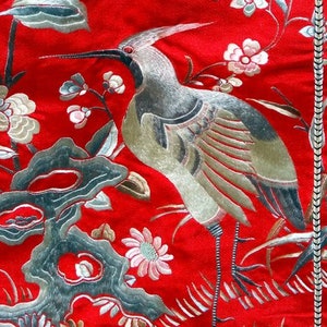 K&ouml;nnte beinhalten: Ein roter Stoffhintergrund mit silberner und goldener Stickerei eines Vogels und Blumen. Der Vogel ist ein Kranich mit langem Hals und langem Schnabel. Die Blumen sind stilisiert und haben verschiedene Formen und Gr&ouml;&szlig;en.
