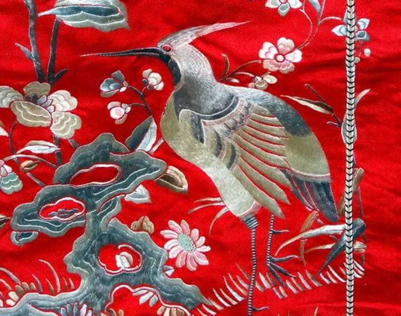 K&ouml;nnte beinhalten: Ein roter Stoffhintergrund mit silberner und goldener Stickerei eines Vogels und Blumen. Der Vogel ist ein Kranich mit langem Hals und langem Schnabel. Die Blumen sind stilisiert und haben verschiedene Formen und Gr&ouml;&szlig;en.