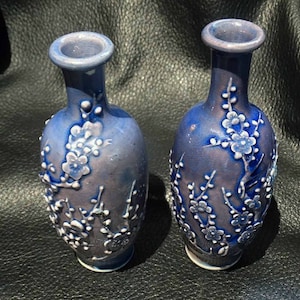Könnte beinhalten: Zwei kleine blaue und weiße Keramikvasen mit einem Blumendesign. Die Vasen sind mit weißen Blumen und Zweigen auf blauem Hintergrund verziert.