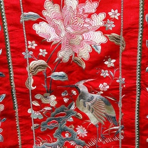 K&ouml;nnte beinhalten: Roter Stoff mit gestickten Blumen- und Vogelmotiven. Der Stoff zeigt eine gro&szlig;e wei&szlig;e Blume mit rosa Akzenten, kleinere wei&szlig;e Blumen und einen Vogel mit langem Hals und braunen und wei&szlig;en Federn.