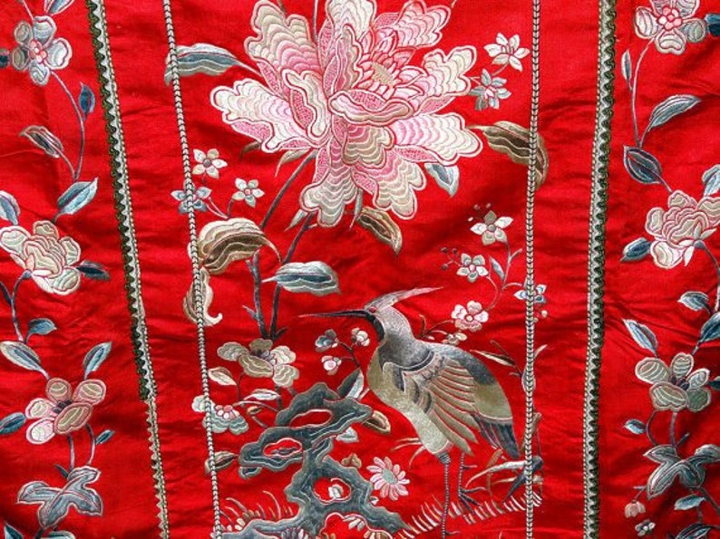 K&ouml;nnte beinhalten: Roter Stoff mit gestickten Blumen- und Vogelmotiven. Der Stoff zeigt eine gro&szlig;e wei&szlig;e Blume mit rosa Akzenten, kleinere wei&szlig;e Blumen und einen Vogel mit langem Hals und braunen und wei&szlig;en Federn.