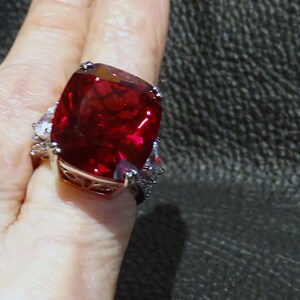 May include: A large, silver-toned ring featuring a vibrant, square-cut, deep red gemstone as the centerpiece. The ring is accented with smaller, clear stones on either side of the main stone, creating a striking contrast. The ring's band has an ornate design.