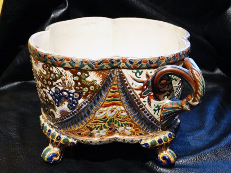 K&ouml;nnte beinhalten: Ein wei&szlig;er Keramiktopf mit einem dekorativen Blumendesign in Blaut&ouml;nen, Gr&uuml;n, Rot und Gold. Der Topf hat drei Beine und einen Griff an einer Seite.