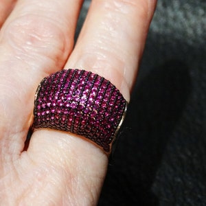 May include: A wide, silver band ring with a deep pink, faceted gemstone setting. The ring is set with small, round stones that cover the entire surface of the band.