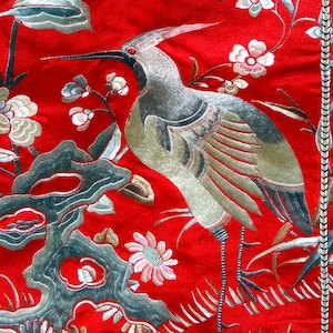 K&ouml;nnte beinhalten: Nahaufnahme eines roten Stoffes mit silberner Stickerei eines Vogels und Blumen. Der Vogel ist ein Kranich mit ausgebreiteten Fl&uuml;geln. Die Blumen bl&uuml;hen und haben zarte Bl&uuml;tenbl&auml;tter.