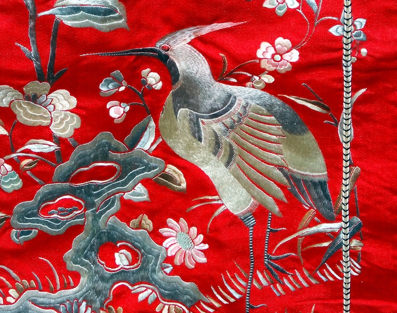 K&ouml;nnte beinhalten: Nahaufnahme eines roten Stoffes mit silberner Stickerei eines Vogels und Blumen. Der Vogel ist ein Kranich mit ausgebreiteten Fl&uuml;geln. Die Blumen bl&uuml;hen und haben zarte Bl&uuml;tenbl&auml;tter.