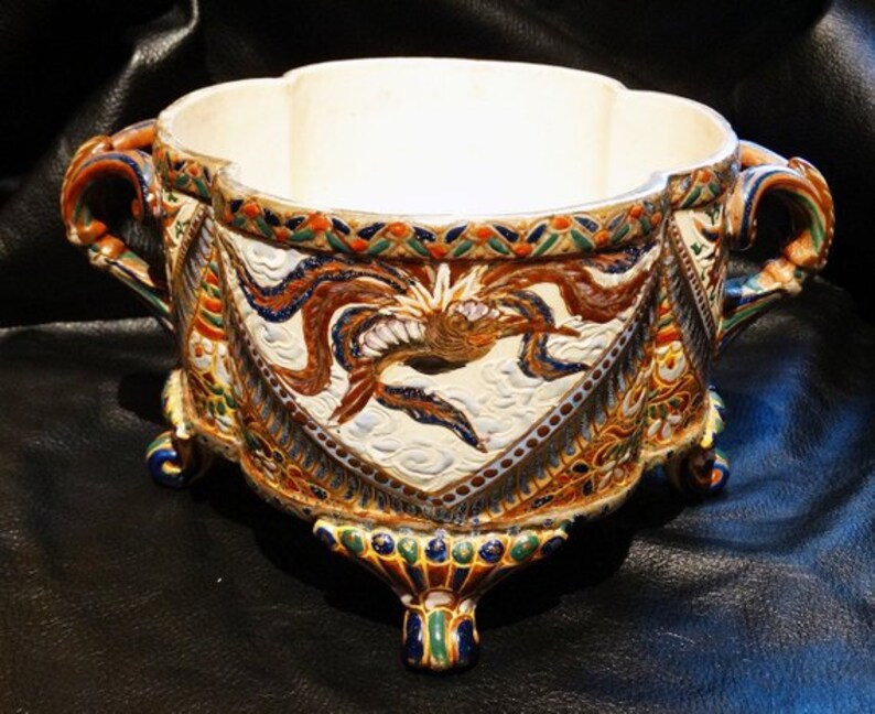 K&ouml;nnte beinhalten: Eine gro&szlig;e, wei&szlig;e Keramikschale mit einem dekorativen Design aus Drachen und Wolken in Blaut&ouml;nen, Gr&uuml;n, Gold und Braun. Die Schale hat drei F&uuml;&szlig;e und zwei Griffe.
