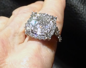 May include: A dazzling silver ring with a square, multi-stone cluster design. The ring features numerous small, sparkling stones set closely together, creating a brilliant, eye-catching effect. The band is also adorned with smaller stones.
