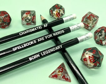 Puede incluir: Lápices negros con texto blanco que dice "CHARISMATIC AF", "SPELLBOOKS ARE FOR NERDS" y "BORN LEGENDARY" están dispuestos con varios dados. Los dados son transparentes con números rojos y un diseño de calavera.