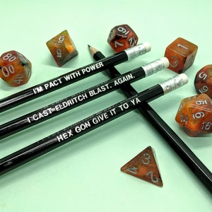 May include: Three black pencils with white text on them. The text on the pencils reads "I'm Pact With Power", "I Cast Eldritch Blast. Again.", and "Hex Gon Give It To Ya". There are several orange and black dice scattered around the pencils.