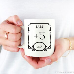 K&ouml;nnte beinhalten: Wei&szlig;e Keramiktasse mit schwarzem Design. Das Design zeigt einen dekorativen Rahmen mit den Worten "SASS +5 20" darin.