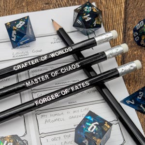 Puede incluir: Tres lápices negros con detalles plateados y letras blancas. Los lápices están etiquetados como "Crafter of Worlds", "Master of Chaos" y "Forger of Fates". Los lápices están sobre una hoja de papel blanca con un dado negro y azul en el fondo.