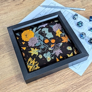 May include: A black wooden dice tray with a patterned fabric insert. The fabric displays a floral design with butterflies in orange, yellow, purple, and green on a black backdrop. Dice and a pencil are visible.