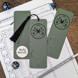 Könnte beinhalten: Salbeigrüne Lesezeichen mit schwarzer Quaste und einem kreisförmigen Design mit einem Hirschkopf und dem Text "I'M A FORCE OF NATURE". Ein Bleistift mit dem Text "ROLL HIGH OR DIE TRYING" ist ebenfalls vorhanden. Die Lesezeichen liegen neben einem Dungeons & Dragons Charakterbogen.