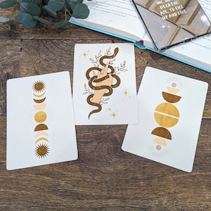 Könnte beinhalten: Drei druckbare Karten mit minimalistischer Ästhetik, die verschiedene Himmelsmotive zeigen. Die erste Karte zeigt eine braune Schlange, die sich um eine Hand mit goldenen Sternen und Blättern windet. Die zweite Karte zeigt einen vertikalen Stapel aus braunen und goldenen Monden und Sonnen. Die dritte Karte zeigt einen vertikalen Stapel aus braunen und goldenen Halbkreisen.