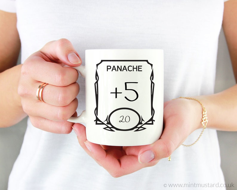 K&ouml;nnte beinhalten: Wei&szlig;e Keramiktasse mit einem schwarzen Design mit den Worten "Panache +5 20" in einem dekorativen Rahmen.