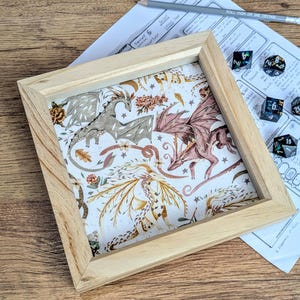 May include: A wooden framed tray with a dragon design. The artwork features dragons in shades of brown and pink, set against a white background with floral and star accents. Dice and a pencil are visible.