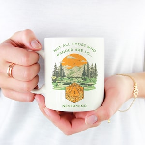 Könnte beinhalten: Eine weiße Keramiktasse mit einer grünen, orangefarbenen und braunen Illustration eines Sonnenuntergangs über Bergen und einem Wald. Die Tasse trägt den Text "Not all those who wander are lo... Nevermind" und einen stilisierten orangefarbenen und braunen Würfel mit der Zahl 1 darauf.