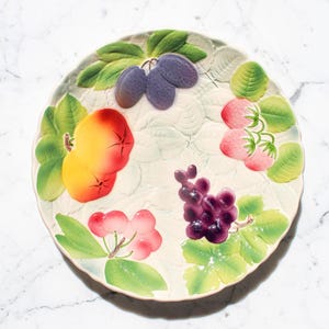 Pode incluir: Um prato decorativo de cerâmica com um design de frutas em relevo. O prato apresenta ameixas, pêssegos, uvas e morangos em cores vibrantes. Folhas verdes e um fundo texturizado completam o design.