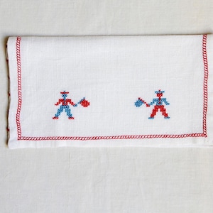 May include: White linen napkin with red stitched border and two embroidered figures in red and blue. The figures appear to be holding objects. The napkin is folded and has a simple, classic design.