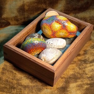 May include: A wooden tray contains two painted stones with colorful dot patterns, two seashells, and a wooden disc. The stones feature vibrant hues of yellow, orange, blue, and purple. The tray is set on a textured, brown surface.