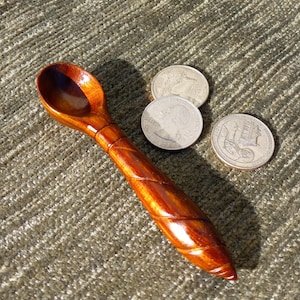 May include: A handcrafted wooden spoon with a polished, reddish-brown finish. The spoon has a deep bowl and a handle with a carved, spiral design. Three US quarter coins are next to the spoon.