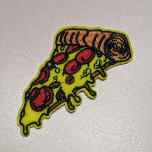 Peut inclure: Un écusson brodé en forme de part de pizza avec une croûte jaune, des cercles de pepperoni rouges et des garnitures vertes. Le motif présente une croûte brune épaisse et texturée sur le dessus avec des coutures noires soulignant la forme.