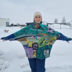 以下が含まれることがあります： 風景デザインのカラフルなフェルトウールポンチョを着た女性。ポンチョは青、緑、紫、黄色の色合いで、ベージュの帽子とブルージーンズが特徴です。背景は雪景色です。