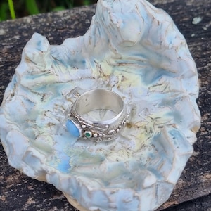 May include: A silver ring with a light blue gemstone and a small green stone, displayed on a handmade ceramic dish. The dish is white with blue and yellow accents, resembling a small, sculpted landscape. The ring has ornate detailing.