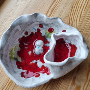 May include: White ceramic bowl with a red glaze and a small, separate compartment. The bowl has a unique, organic shape and is decorated with red and green speckles.
