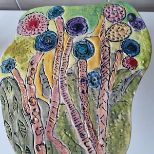 以下が含まれることがあります： 緑色の背景にカラフルな花が描かれたセラミック製の壁飾り。花はピンク、パープル、ブルー、イエロー、レッドの色合いで描かれています。作品はテクスチャのある表面で、葉の形をしています。