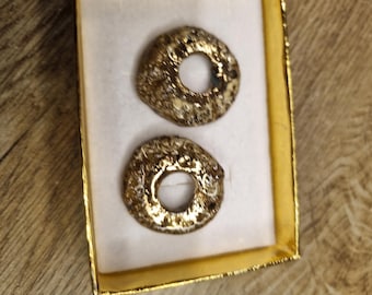 May include: Two circular, metallic gold-toned objects with a textured, uneven surface and a central hole, presented inside a rectangular box with a white interior lining and a metallic gold-colored frame.
