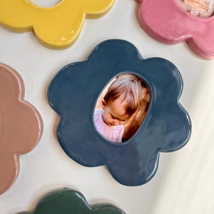 May include: A collection of flower-shaped ceramic photo frames in various colors, including yellow, pink, blue, and green. Each frame holds a small photograph of a child. The frames have a glossy finish and are arranged on a white surface.