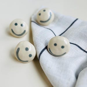 May include: Four off-white ceramic smiley face napkin rings with gray details. One ring is on a white and gray striped napkin. The rings have a smooth, rounded shape and a simple, cheerful design.