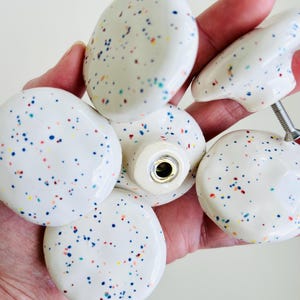 May include: A collection of five round ceramic cabinet knobs with a glossy off-white finish and a multicolored speckled pattern featuring small dots of blue, red, yellow, and green. Each knob has a central metal screw attachment on the back.