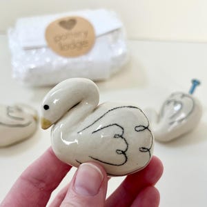May include: Three ceramic drawer knobs shaped like swans with a glossy off-white finish, black line details on the wings, and a yellow beak. Each swan knob features a blue metal screw protruding from the back for installation purposes.