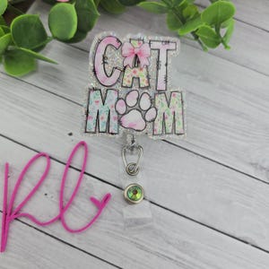 May include: A clear badge reel with the words "CAT MOM" in pink and floral print. The design includes a paw print and a pink bow. The reel has a silver clip and a decorative iridescent gem.