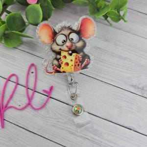 May include: A glittery badge reel featuring a cartoon mouse holding a block of cheese. The mouse has large eyes, pink ears, and a long tail. The reel has a clear plastic strap and a decorative button.